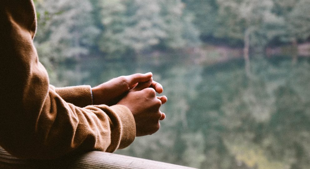 Close-up photo of the hands of a person leaving over a deck overlooking a lake surrounded by trees.