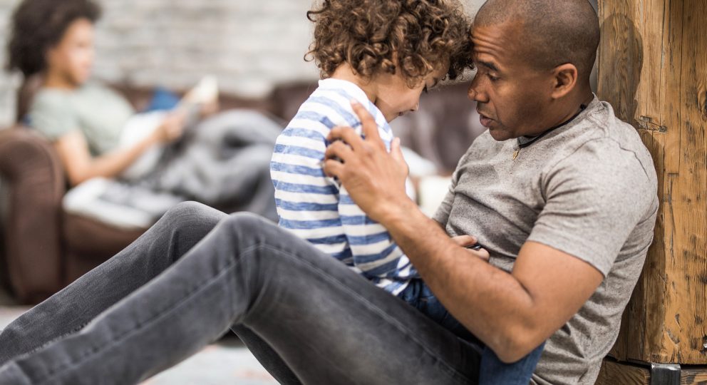 A photo of an African-American father sitting with his son in his lap. He is talking to his son, who looks away from his father.